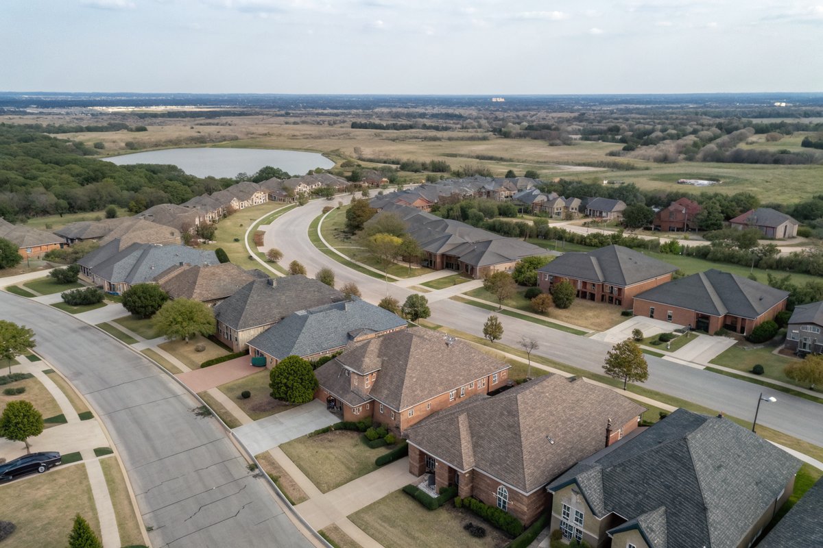 Aerial view of a suburb of brick homes with gray and brown shingle roofs around a small pond