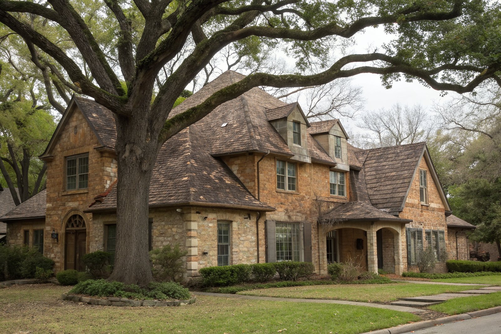 A two-story brick and stone home with a weathered brown shingle roof behind a large pecan tree